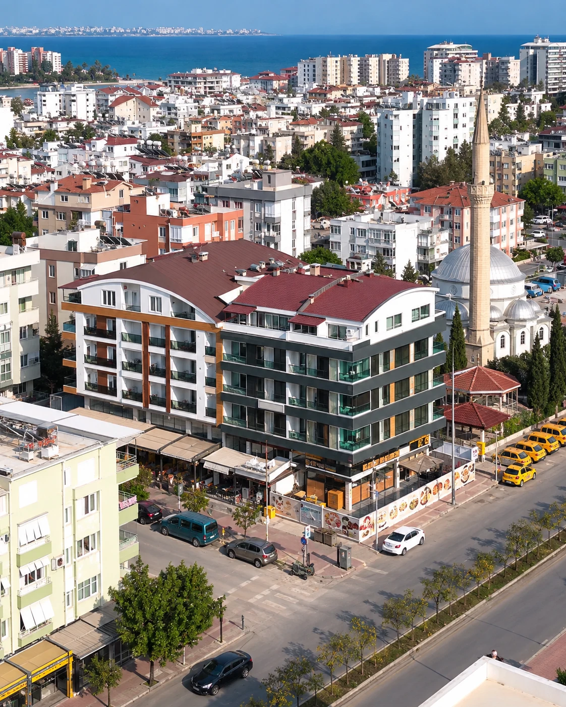 7 Lezzet Lokantası’nın bulunduğu koyu cepheli köşe bina; sağında cami ve taksi durağı.
